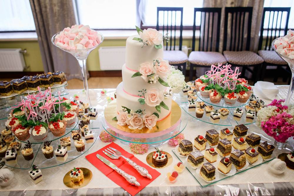 A three tier wedding cake surrounded by smaller cupcakes and sweets